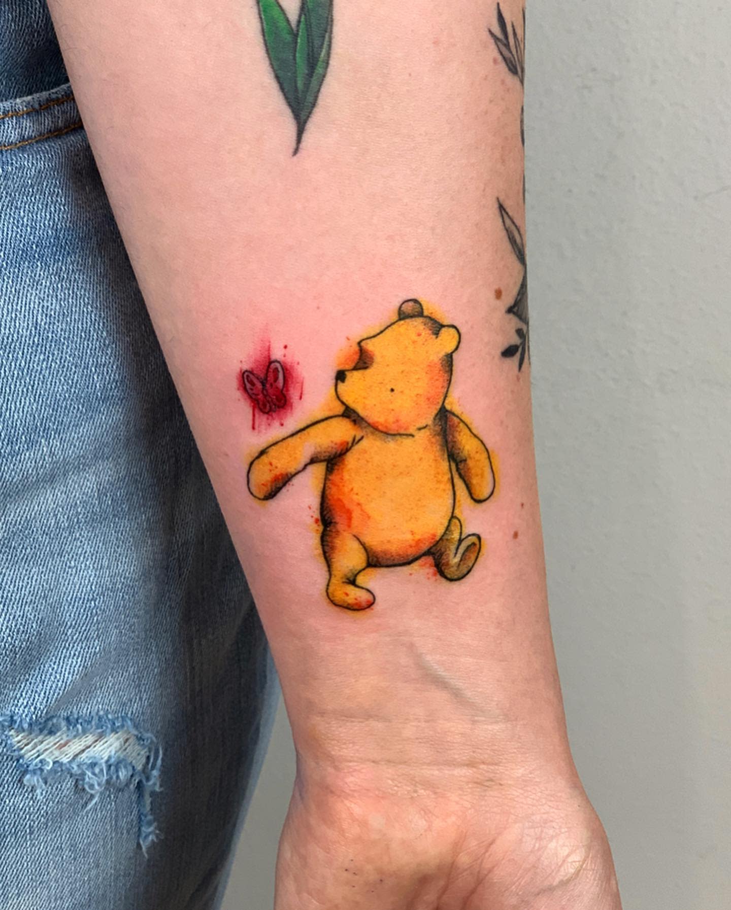 Watercolor tattoo of Winnie the Pooh looking at a small red butterfly on a forearm